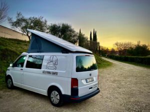 Weißer VW California Campervan mit ausgeklapptem Aufstelldach bei Sonnenuntergang auf einem Schotterweg, umgeben von Bäumen – Köllefornia Camper Logo an der Fahrzeugseite.